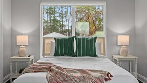 Une chambre avec un grand lit, deux tables de chevet, une fenêtre donnant sur des arbres et un palmier, et des coussins à rayures vertes.