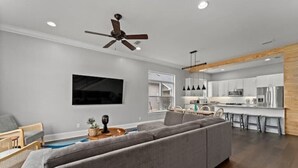 Un salon moderne avec un canapé d’angle gris, un téléviseur à écran plat fixé au mur, un ventilateur de plafond et un mur décoratif en bois.