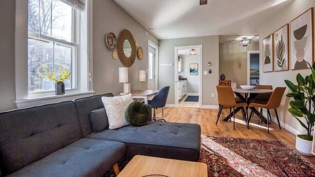 Ein modernes Wohnzimmer mit einem dunkelgrauen Ecksofa, einem Holztisch und einem gemusterten Teppich. Es gibt einen Essbereich mit einem runden Tisch und Stühlen, sowie eine Küche im Hintergrund.