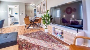Ein modernes Wohnzimmer mit großem Flachbildfernseher, einem Essbereich mit Tisch und Stühlen sowie einer gemütlichen Sitzecke mit gemustertem Teppich.
