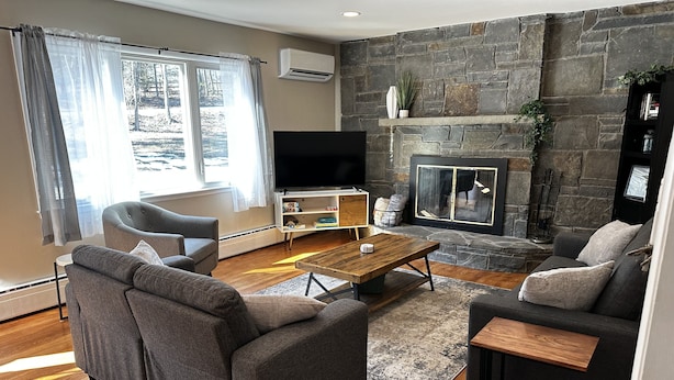 A modern living room with a stone fireplace, grey sofas, a wooden coffee table, and a flat-screen TV. - Modern Country Farmhouse, Walking Distance to Lake (Averill Park)