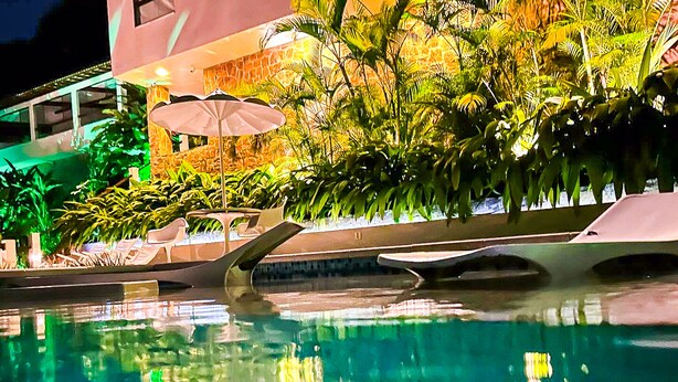 A swimming pool with a reflection of palm trees and a building with a stone facade. - Oasis Beach House (Angra dos Reis)