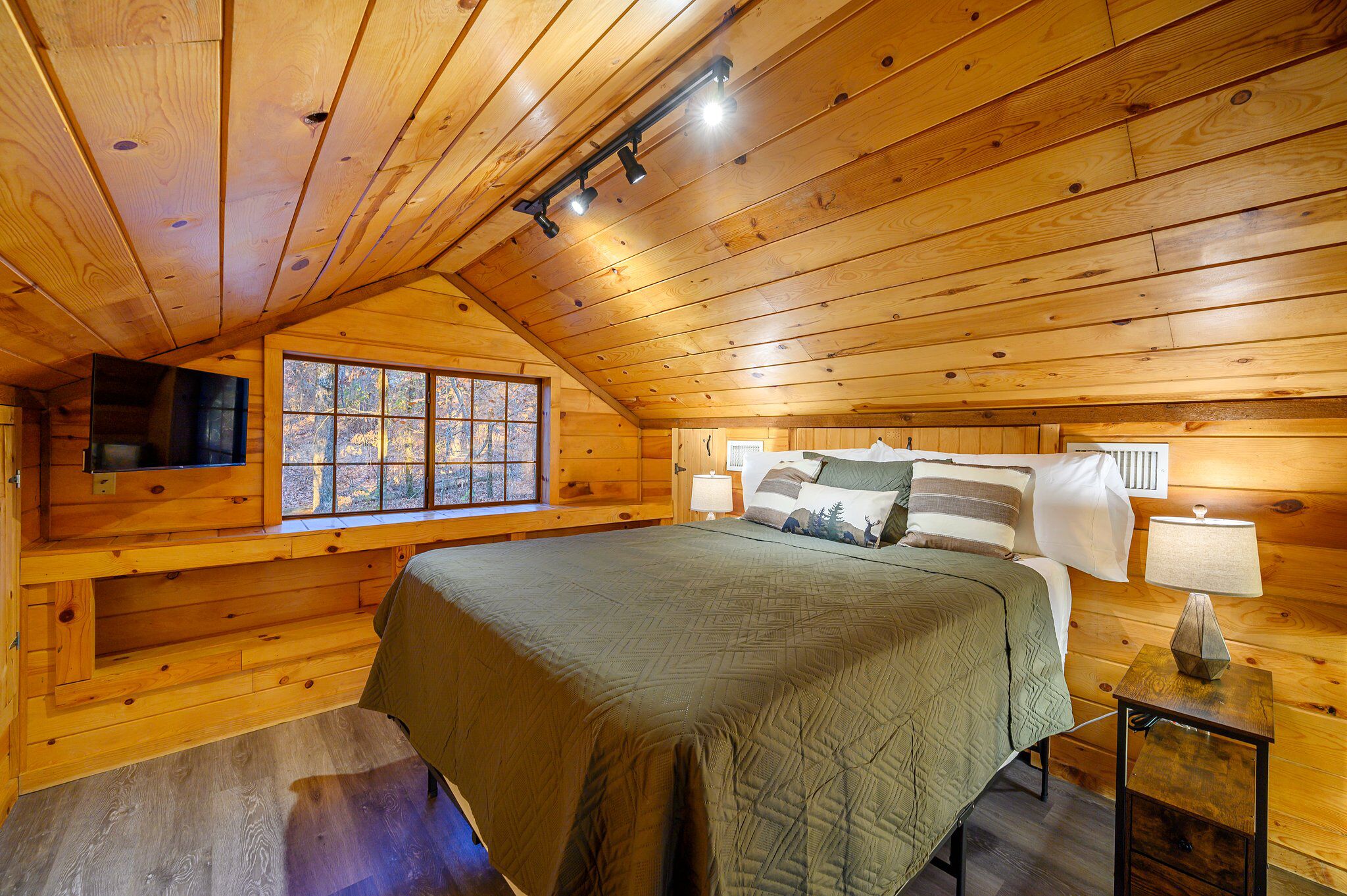 A cozy cabin bedroom with a large bed, a TV mounted on the wall, and a window with a view of trees.