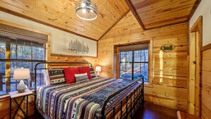 Un dormitorio de cabaña de madera con cama, dos lámparas de noche, una ventana con vista a árboles y una lámpara de techo.