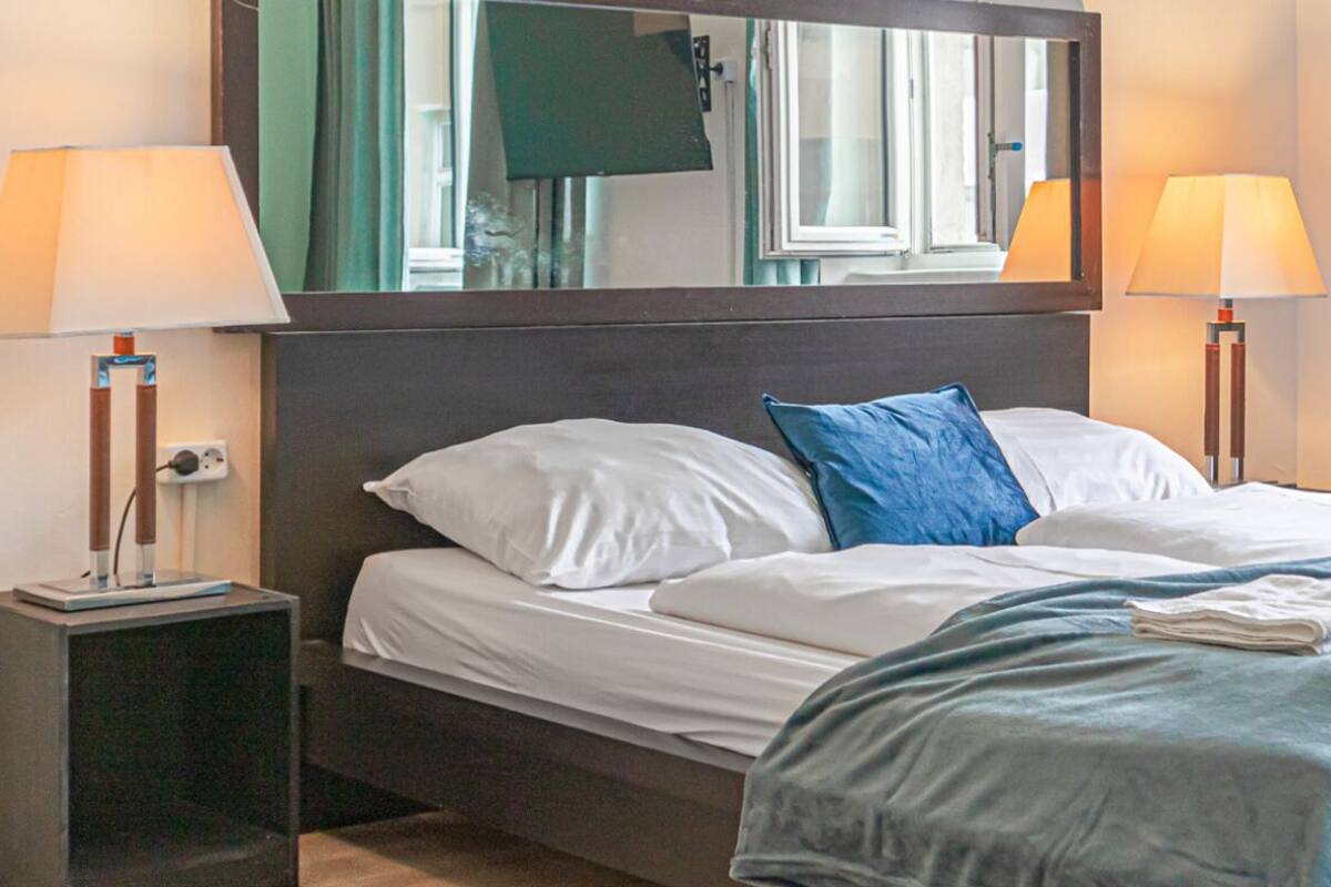 A neatly made bed with white and blue bedding, two bedside lamps, a mirror above the bed, and a window with curtains.