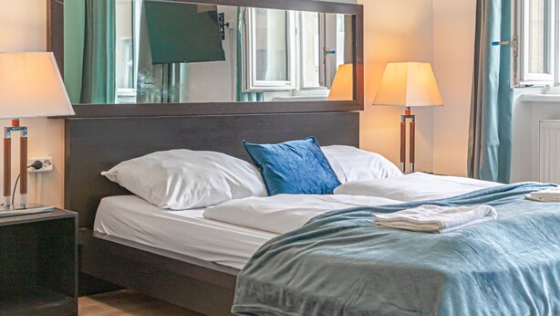Ein ordentlich bezogenes Bett mit weißer und blauer Bettwäsche, zwei Nachttische mit Lampen, ein Spiegel über dem Bett und ein Fenster mit Vorhängen.
