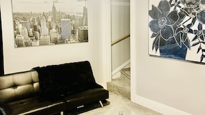 Un salon avec un canapé en cuir noir, une table basse en verre, un ventilateur de plafond et deux tableaux encadrés accrochés aux murs.