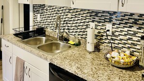 Une cuisine avec des meubles blancs, un plan de travail en granit, un évier à deux bacs, un égouttoir à vaisselle, un porte-essuie-tout et un panier de bananes.