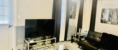 Un salon avec un téléviseur à écran plat posé sur un meuble, une table basse en verre, un canapé en cuir noir et un radiateur.