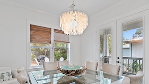 A dining area with a glass table, white chairs, a wooden-framed chandelier, and a view of the outdoors.