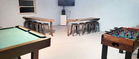 Una habitación con mesa de billar, dos mesas de futbolín y un televisor montado en la pared.