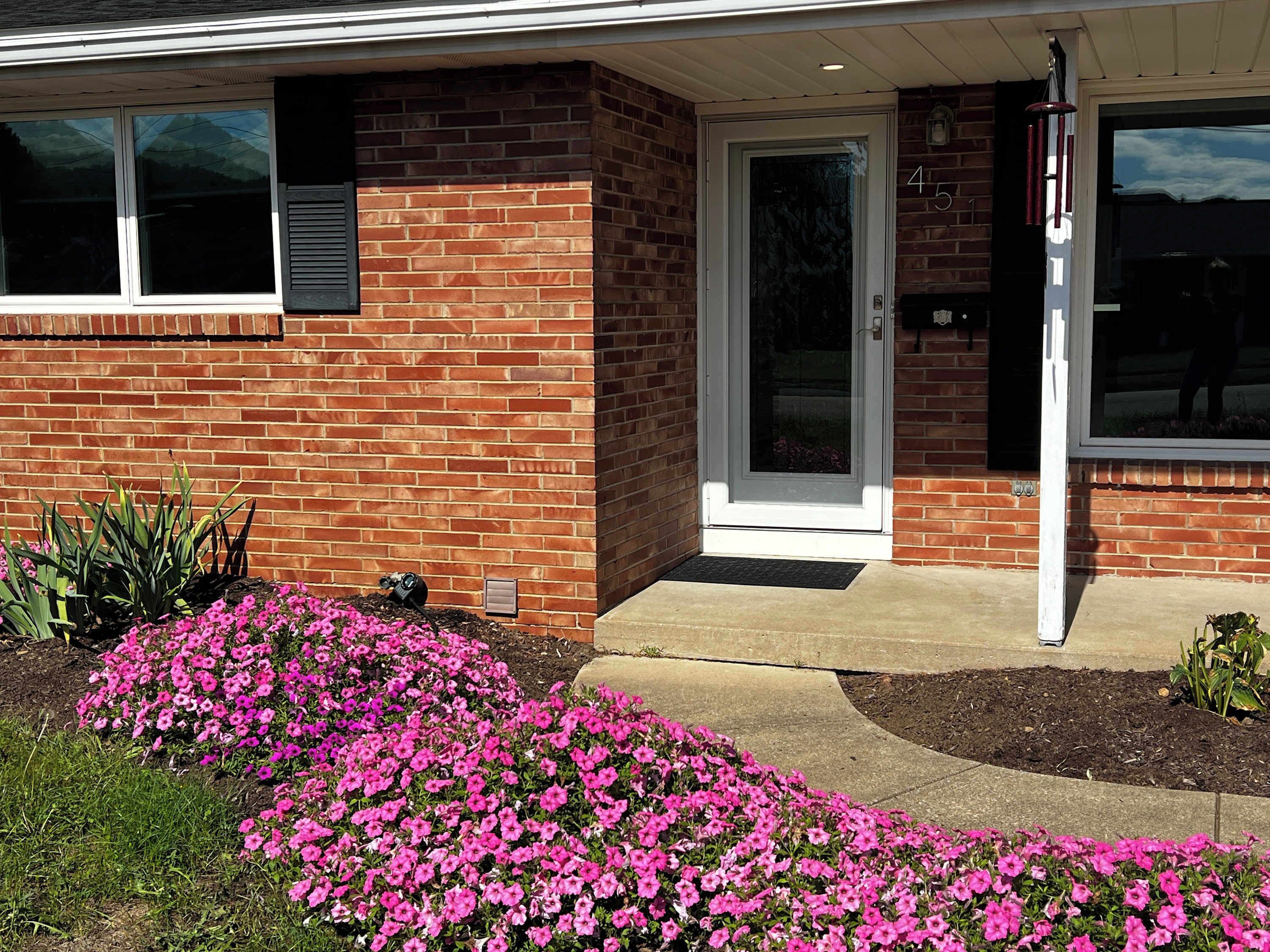 A brick house with a white door, a small porch, and pink flowers in the front yard.