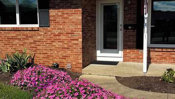 A brick house with a white door, a small porch, and pink flowers in the front yard.