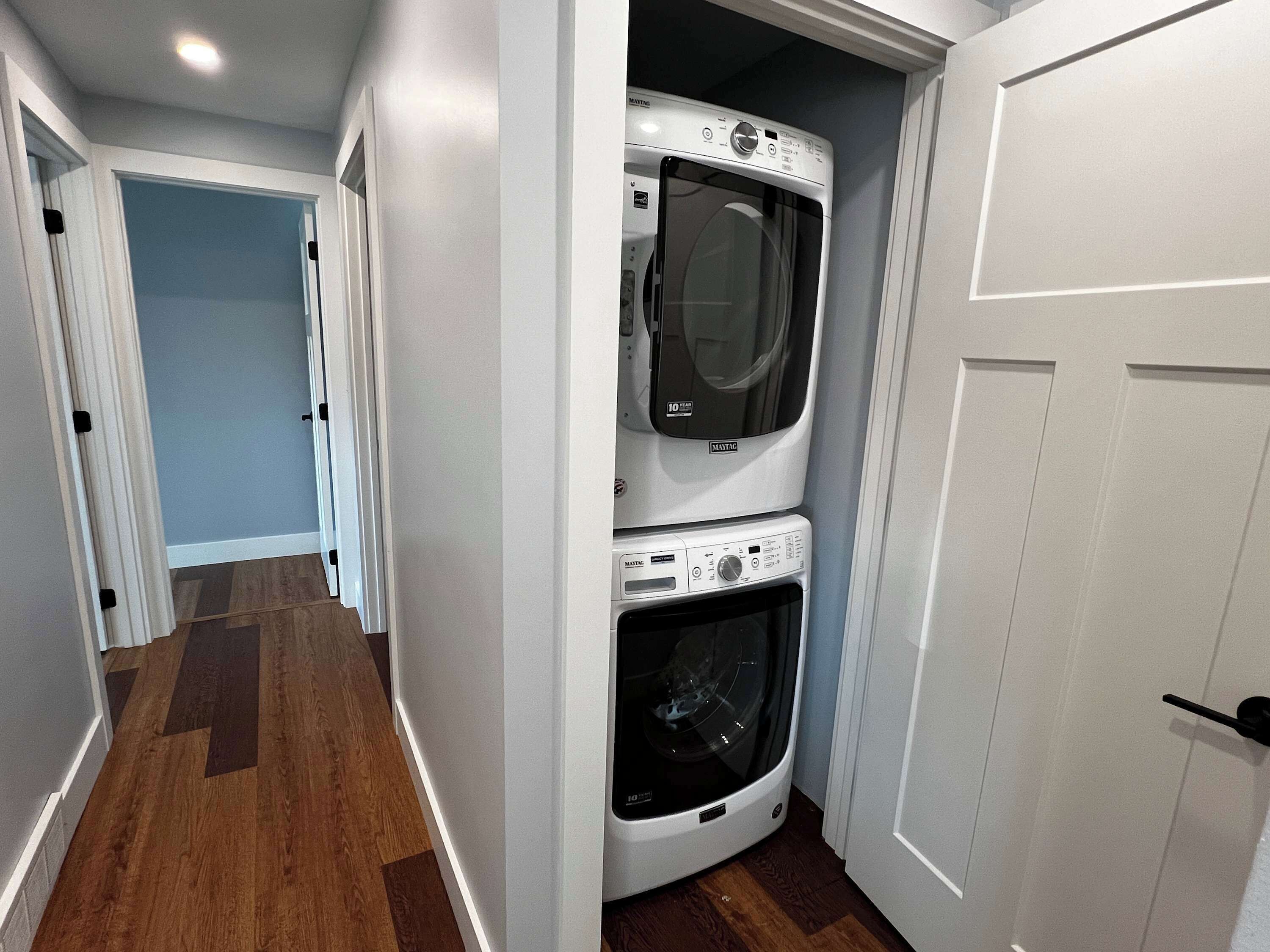 A laundry room with a stacked washer and dryer, wooden flooring, and a door leading to another room.