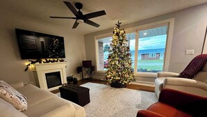 Een woonkamer met een open haard, een kerstboom, een plafondventilator en een aan de muur bevestigde televisie.
