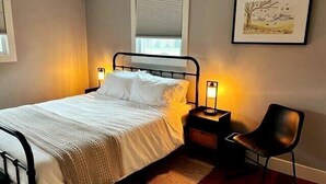 Een net opgemaakt bed met witte lakens, twee bedlampen, een stoel en een lijst met een foto aan de muur.