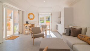 A spacious living area with a sofa, chairs, a coffee table, and a kitchen area visible through an open door. - Chalet Maria da Assunção T1 (Cascais)