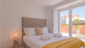 A bedroom with a bed, a bedside table with a lamp, and a view of buildings through a large window. - Chalet Maria da Assunção T1 (Cascais)