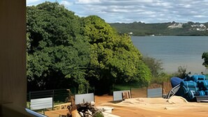 A view from a balcony overlooking a lake, with a forested area and a sandy beach with chairs and a tent. - Brisas do Lago - apt 13 (Brasília)