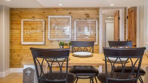 Un comedor con pared de madera, tres letreros enmarcados y cuatro sillas negras alrededor de una mesa de madera.