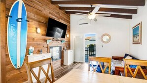 A room with a wooden wall, a surfboard, a TV, a ceiling fan, a dining table, and a bed.