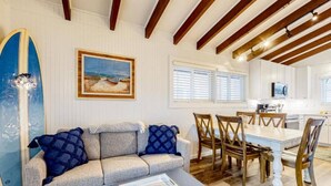 A cozy living area with a sofa, a coffee table, and a surfboard leaning against the wall. There is a dining table with chairs and a kitchen in the background.