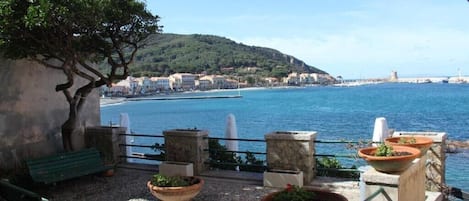 A terrace with potted plants, a bench, and a view of the sea and buildings.