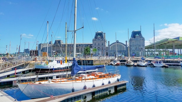 A marina with boats docked, a large building with a distinctive arched entrance, and a bridge connecting to the dock area.