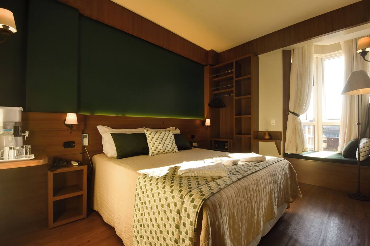 Ein Hotelzimmer mit einem großen Bett, Nachttischen, einer fest installierten Ablage und einem Fenster mit Vorhängen.