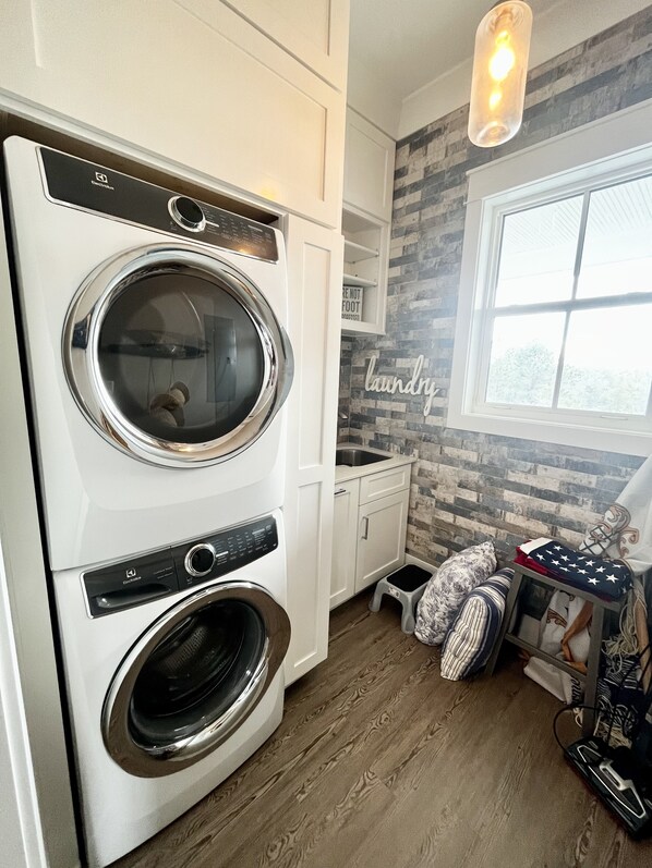 A laundry room with two stacked front-loading washing machines, a small stool, and a laundry basket. - Bayfront! Ultimate sunsets in Bay St. Louis (Bay St. Louis)