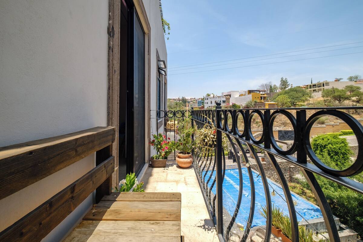 A balcony with a wooden railing, potted plants, and a view of a swimming pool and buildings in the background.