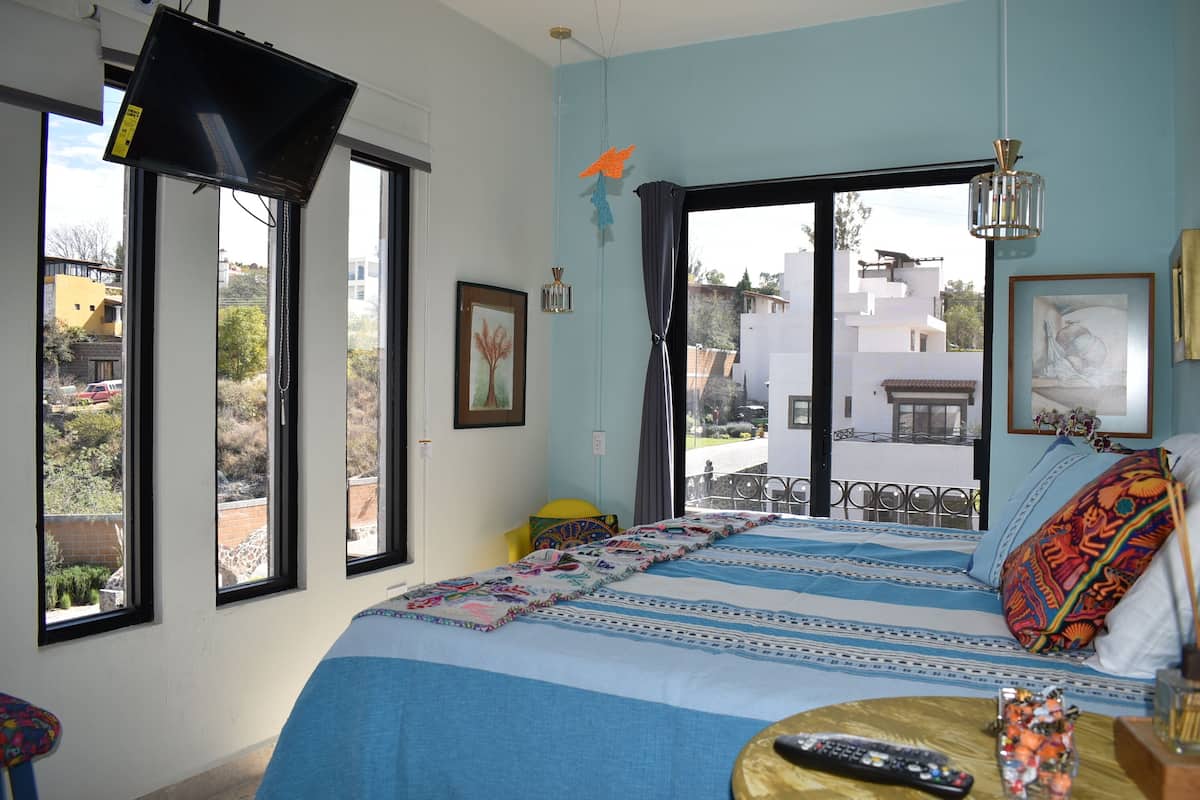 A bedroom with a large bed, a TV mounted on the wall, and a balcony with a view of buildings and trees.