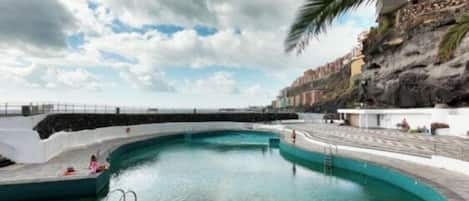 Une piscine incurvée avec une terrasse blanche, un palmier et une falaise rocheuse en arrière-plan.