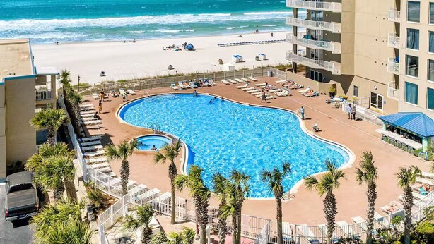 Een hotel met een zwembad naast een strand, omgeven door palmbomen en ligbedden.