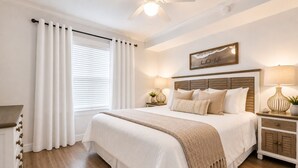 Een slaapkamer met een groot bed, witte gordijnen, een plafondventilator, een nachtkastje met een lamp en een bordje aan de muur.