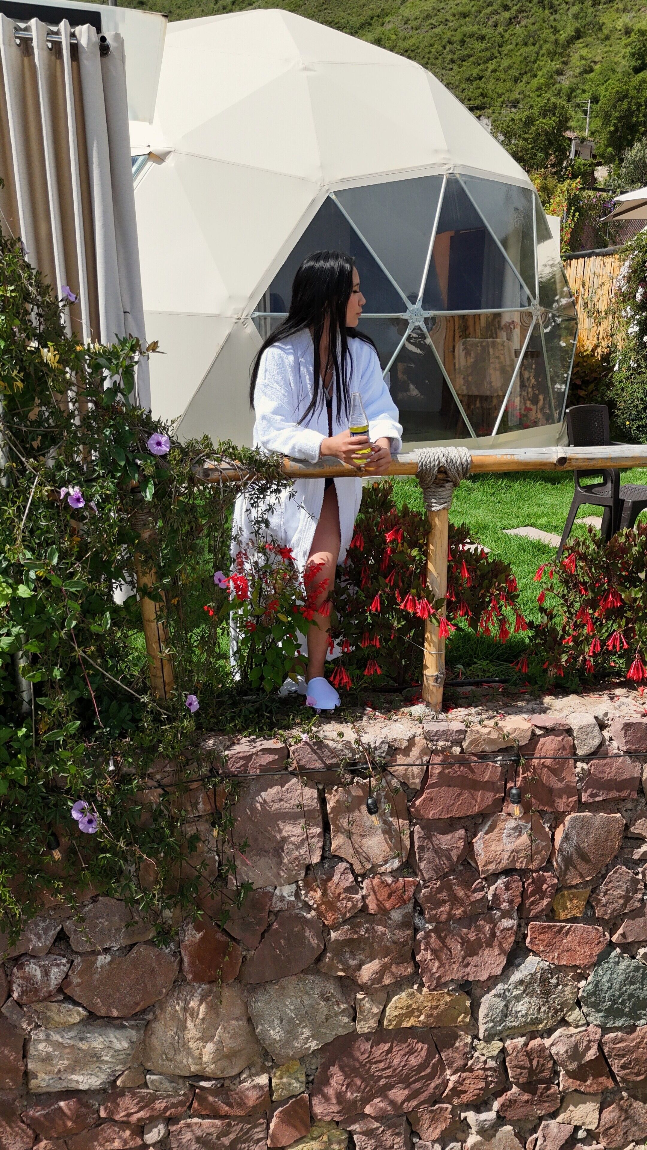 A person sitting on a bench in front of a geodesic dome structure with a stone wall and flowering plants in the foreground.