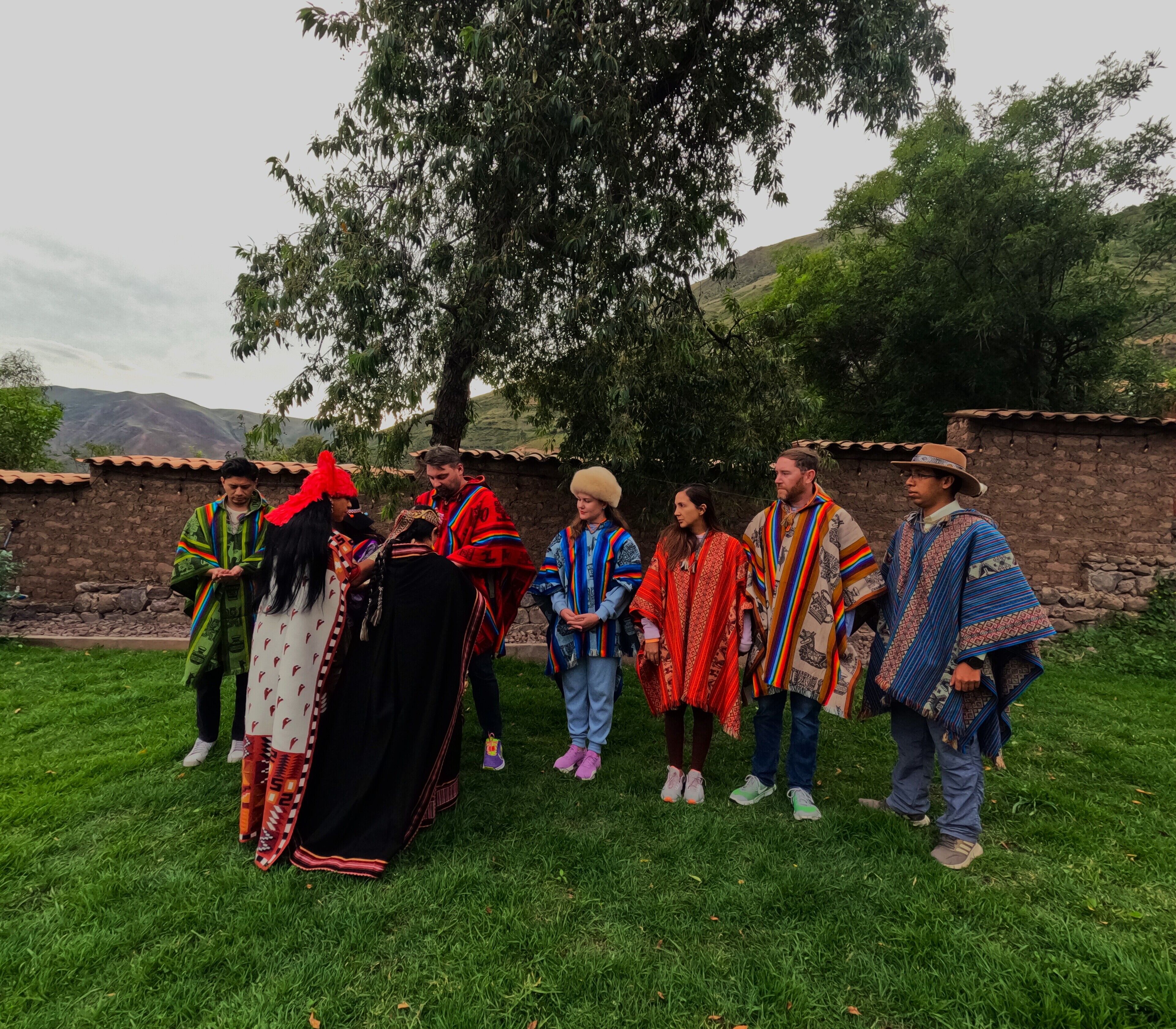 Un groupe de personnes, vêtues de costumes traditionnels, se tient sur une pelouse, avec un mur de pierre et des arbres en arrière-plan.
