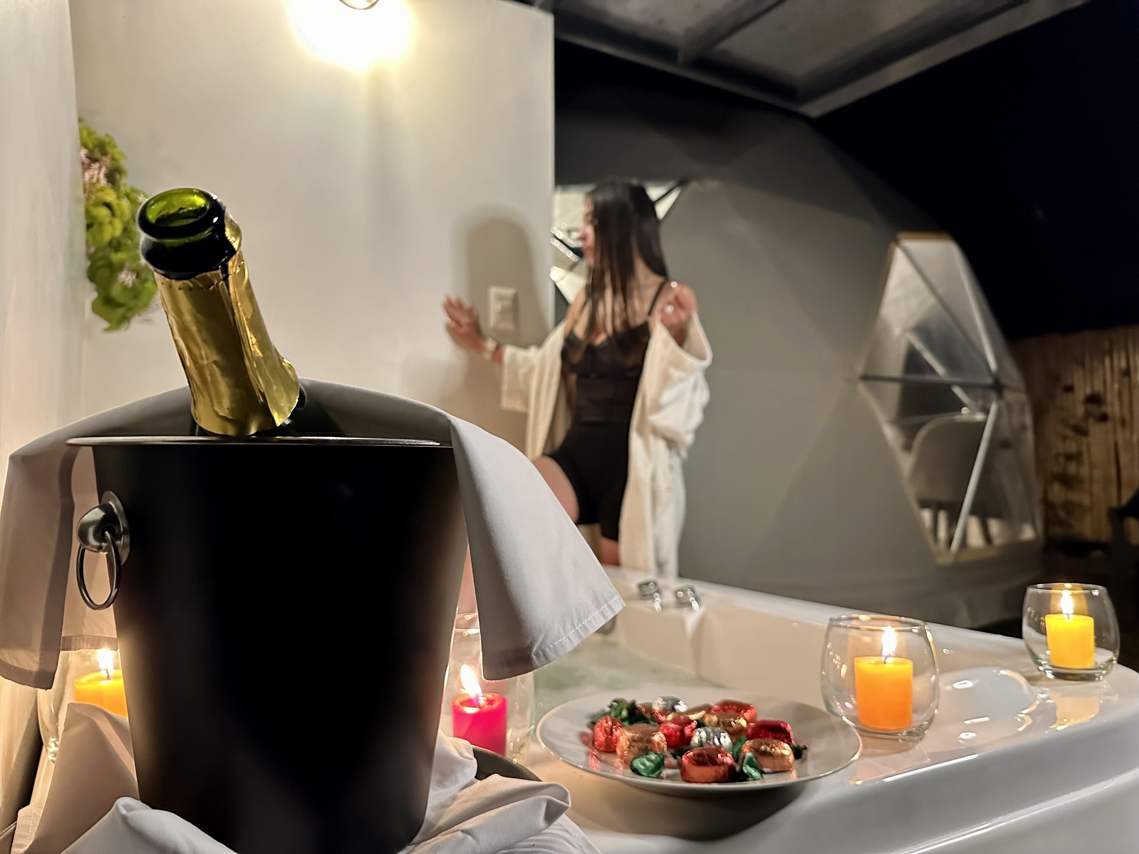 A room with a champagne bottle in a bucket, a candle-lit table with snacks, and a person in the background.