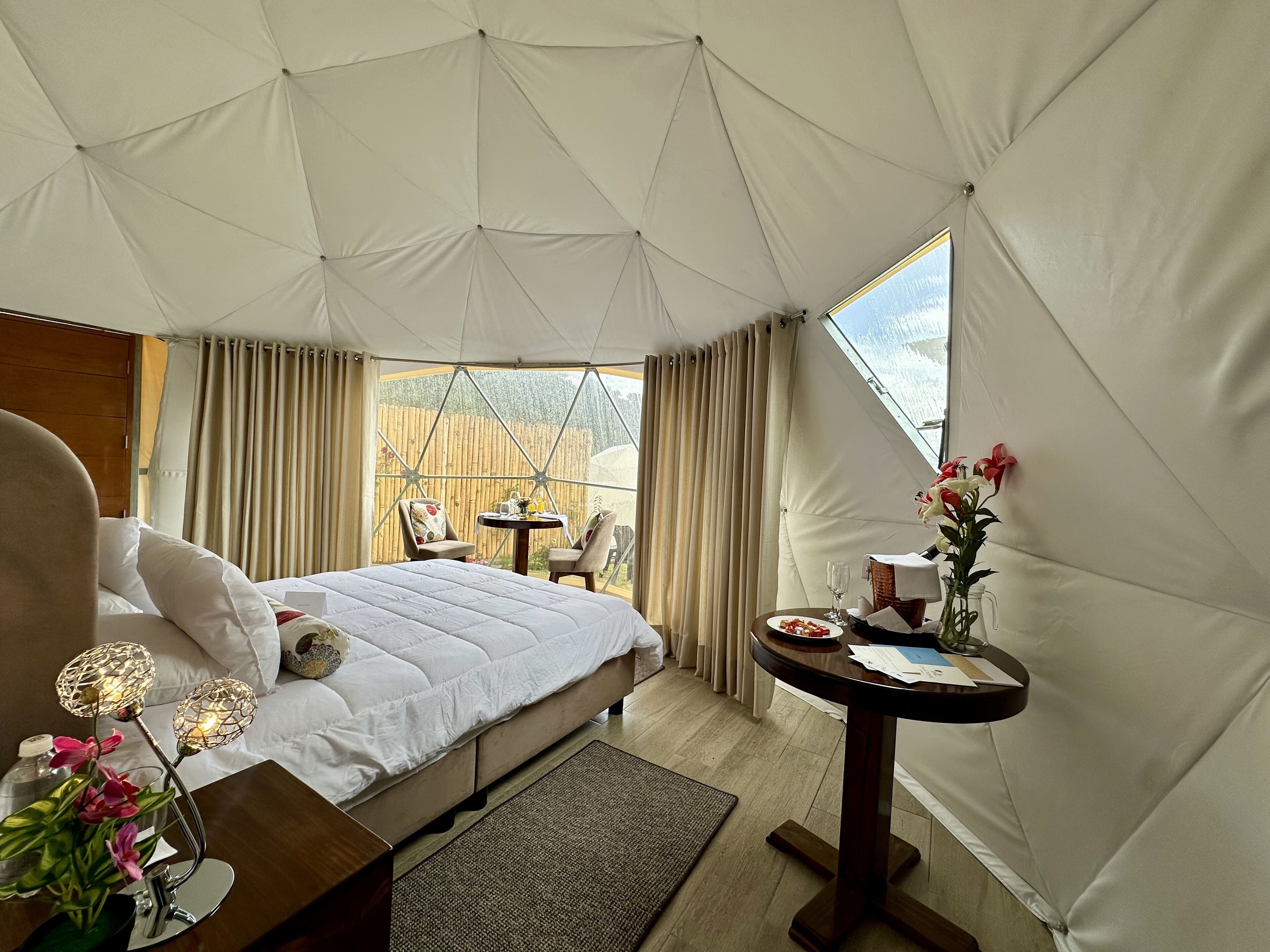 A tent-style room with a bed, a small table with a vase of flowers, and a view of a wooden fence outside.