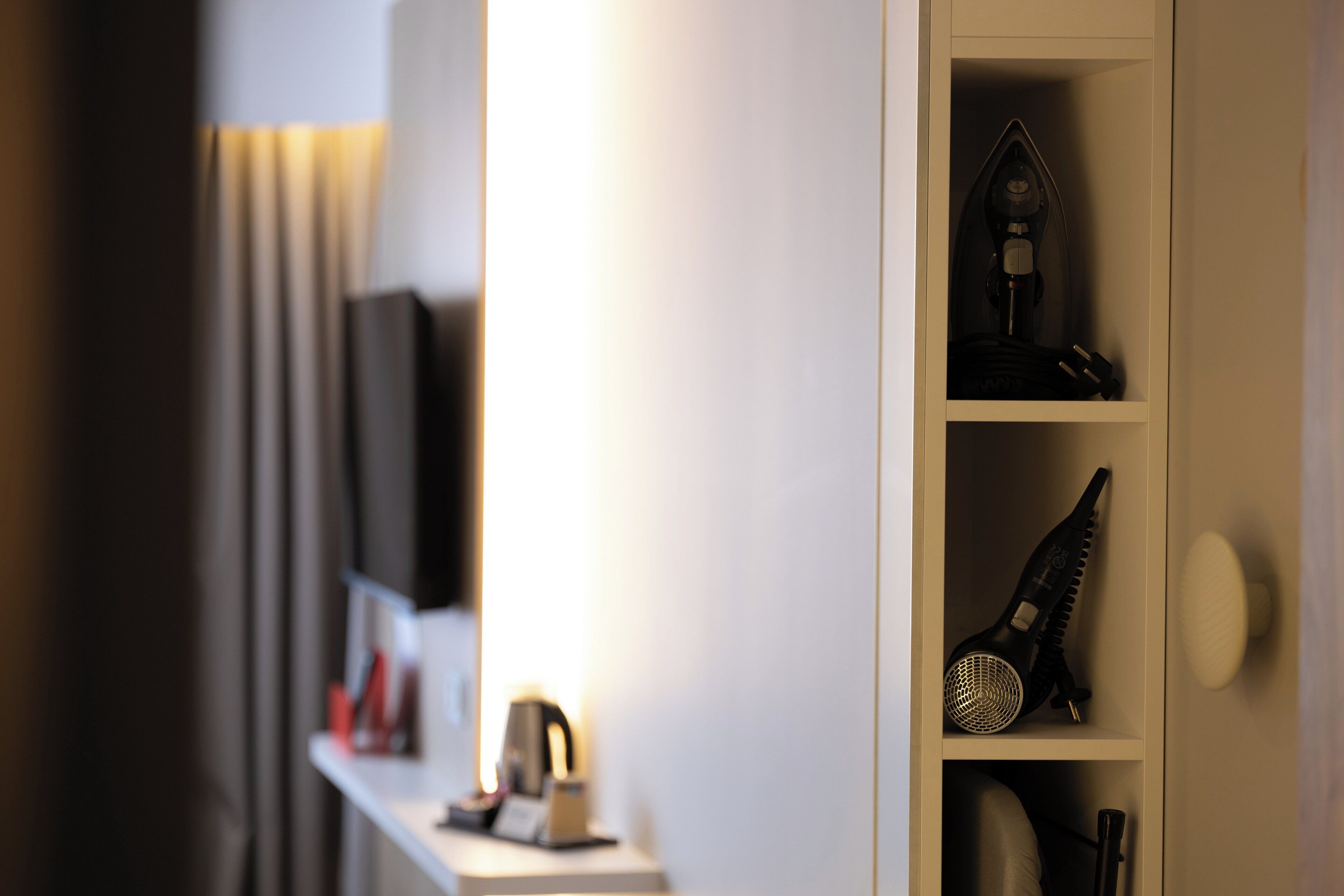 Une chambre d’hôtel avec un mur blanc, une étagère ornée d’un objet décoratif et une table de chevet avec une lampe et une petite horloge.