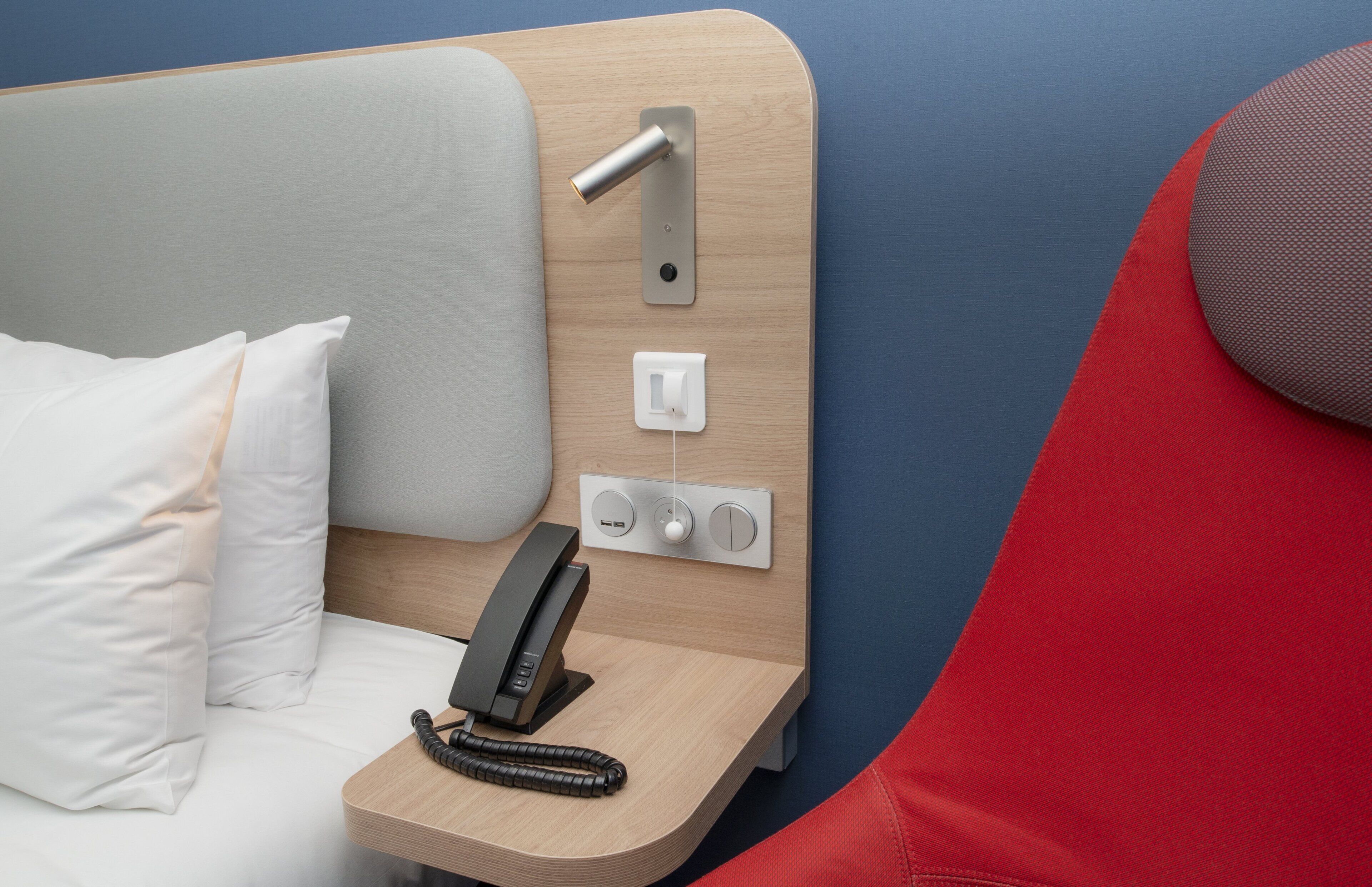 A hotel room with a bed, a bedside telephone, and a control panel with buttons and a switch.
