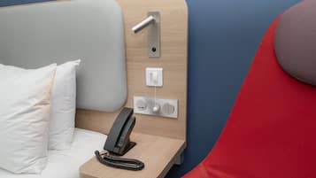 A hotel room with a bed, a bedside telephone, and a control panel with buttons and a switch.