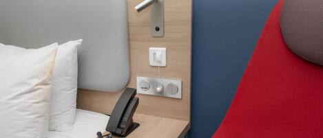 A hotel room with a bed, a bedside telephone, and a control panel with buttons and a switch.