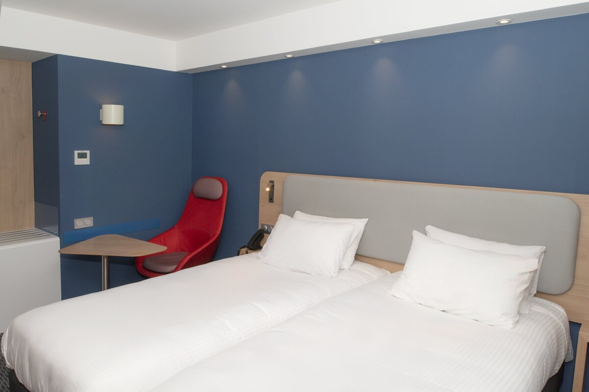 A hotel room with a large bed, a red chair, a small table, and a wall-mounted phone.