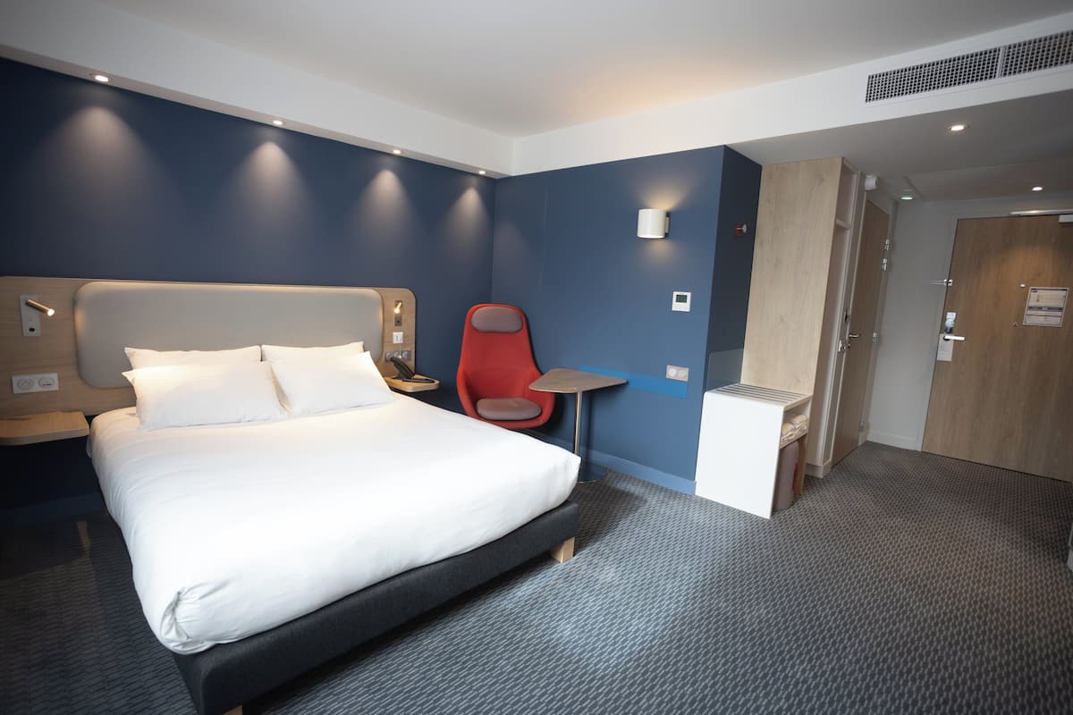 A modern hotel room with a large bed, a red chair, a small table, and a wooden wardrobe.