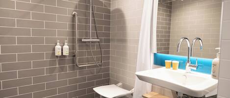 A modern bathroom with grey tiled walls, a white sink, a shower with a handheld showerhead, and two bottles of shampoo on the wall.