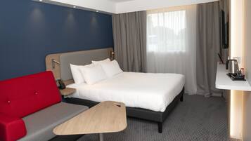 Ein Hotelzimmer mit einem Bett, einem roten Sofa, einem kleinen Holztisch, einem Fernseher und einem Fenster mit Vorhängen.