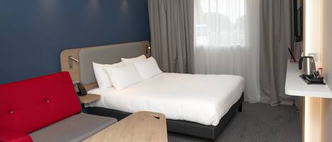 Una habitación de hotel con cama, sofá rojo, una pequeña mesa de madera, televisor y ventana con cortinas.
