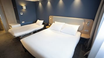 Une chambre d’hôtel avec deux lits simples et un lit plus grand, tous dotés de draps blancs. On y trouve des appliques murales et une petite table de chevet avec une lampe.