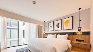 A modern hotel room with a large bed, a wooden nightstand, a wall-mounted lamp, and a large window with curtains. - voco Bali Seminyak by IHG (Keroboken Kelod)